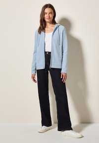 Felpa con zip di colore blu chiaro, realizzata in tessuto morbido, indossata sopra una maglietta bianca, abbinata a jeans scuri e sneakers bianche. Presenta un cappuccio e tasche.
