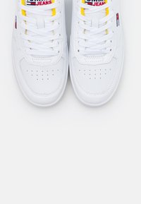 White sneakers with smooth leather upper, round toe, and flat white laces. Features yellow and red fabric accents near the tongue.