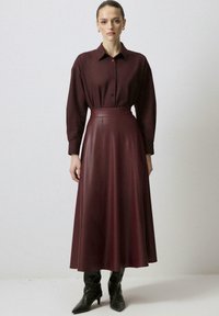 Sieviete ar uzkārtām matiem, ģērbta tumši burgundijas krāsas pogātā krekla un atbilstošā augstās jostas ādas svārkos, kā arī melnās norādītajās zābakos.