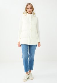 Cremefarbene, gepolsterte Weste mit Kapuze, Druckknöpfen und Seitentaschen. Getragen über einem gerippten Pullover und kombiniert mit hellblauen Jeans und Sneakern.