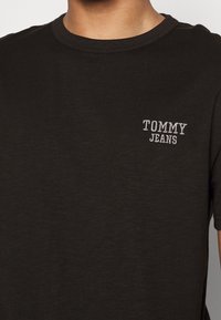 T-shirt en coton noir avec un col rond et des manches courtes. "TOMMY JEANS" brodé en blanc sur le côté gauche de la poitrine. Texture lisse.