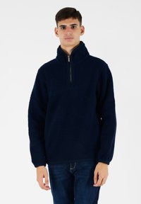 Pullover in pile blu navy con colletto alto, chiusura frontale a metà zip e polsini e orlo leggermente elasticizzati, che offre un aspetto morbido e testurizzato.