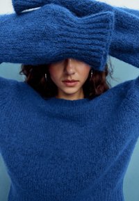 Dlhý rukáv, chlpatý sveter v hlbokej modrej farbe, s rebrovaným golierom a manžetami, doplnený o zodpovedajúce textúrované rukavice zakrývajúce tvár modelu.