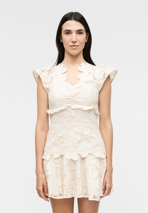 Γυναίκα με μακριά σκούρα μαλλιά φοράει κρεμ δαντελένιο φόρεμα με λουλουδάτα σχέδια, κυματιστές άκρες και βολάν κοντά μανίκια σε απλό φόντο.