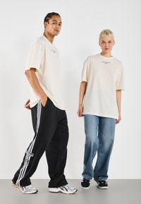 Cream oversized t-shirts, black track pants with stripes, and blue jeans. Sneakers are silver and black. Simple design, relaxed fit.