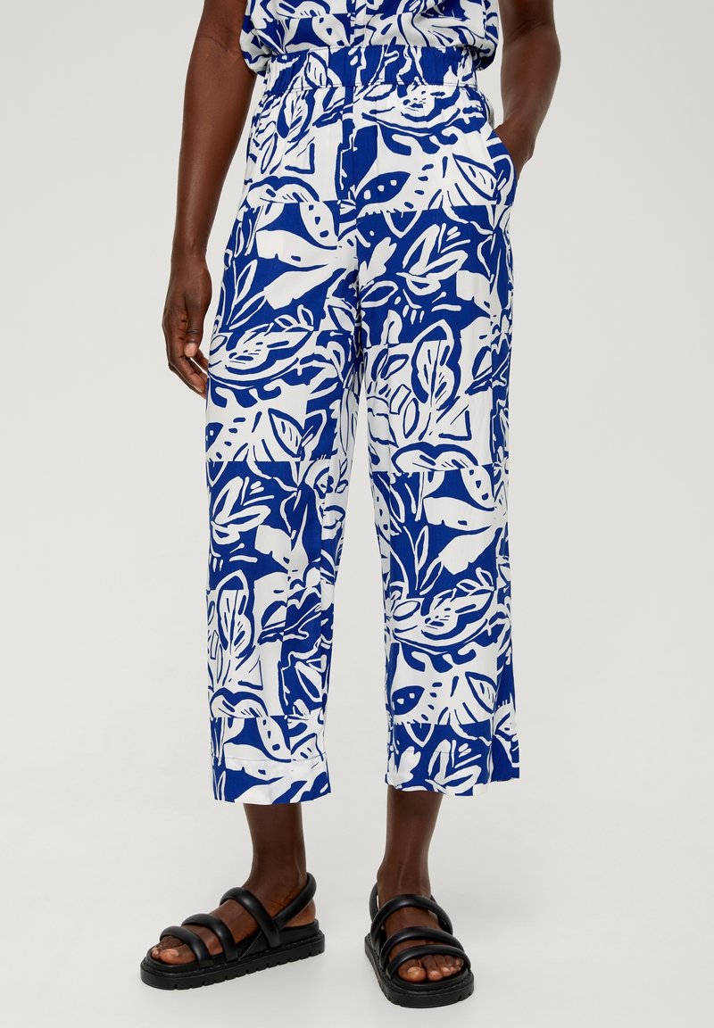 Personne portant un pantalon court à motifs de feuilles bleu et blanc et des sandales noires, avec une main dans la poche, se tenant contre un fond uni.