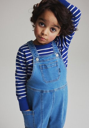 Jong kind met krullend haar, draagt een blauw-wit gestreept shirt met lange mouwen en denim tuinbroek, één hand in de zak, de andere hand raakt het hoofd aan.
