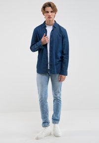 Camisa de mezclilla de botones en azul oscuro con mangas largas, combinada con jeans ajustados de azul claro y zapatillas blancas, contra un fondo neutro.