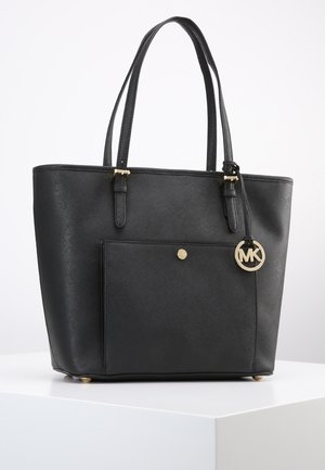 Černá kožená kabelka typu tote s dvojitými rukojeťmi, přední kapsou se zapínáním na druk a zlatým přívěskem s logem MK visícím na boku.