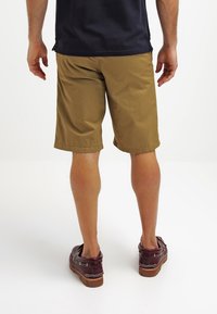 Shorts marron en tissu léger, à mi-cuisse, avec une texture lisse et un ourlet simple. Portés avec des chaussures bateau bordeaux foncé.
