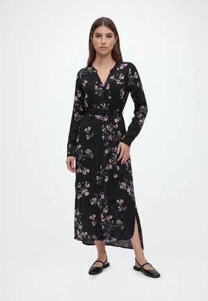 Kvinde iført en sort langærmet blomsterkjole med bælte i taljen og sideslids, kombineret med sorte flade sko, stående foran en hvid baggrund.