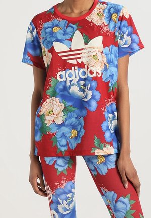 Persoon draagt een rode Adidas-outfit met grote blauwe en witte bloemmotieven; het shirt heeft het witte Adidas-logo op de borst.