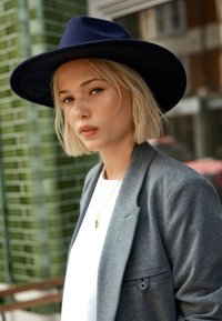Blazer gri peste o bluză albă, împodobită cu un mic colier din aur. Pălărie din fetru de culoare albastru închis cu margine largă. Fundal texturat verde.