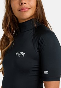 Svart rash guard med kort ärm tillverkad av elastiskt material. Har Billabong-logotypen i vitt på framsidan och UV-skydd-logotypen på ärmen.