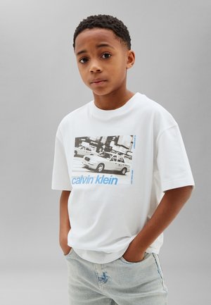 Ung dreng med hænderne i lommerne iført en hvid Calvin Klein t-shirt med et sort-hvidt bytaxa-tryk og lyseblå jeans.