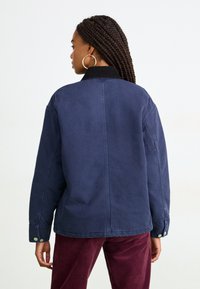 Donna con capelli intrecciati, che indossa orecchini a cerchio grandi, una giacca blu navy con colletto nero e pantaloni di velluto a coste borgogna, in piedi di spalle.