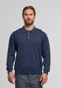 Mann mit Bart und langen Haaren, der ein marineblaues Langarm-Polo-Shirt und graue Hosen trägt, vor einem schlichten hellen Hintergrund.