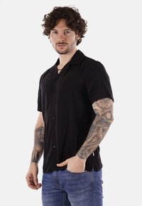 Maglietta nera a maniche corte in tessuto leggero, con apertura frontale a bottoni, colletto a punta e una texture delicata, indossata su jeans blu.