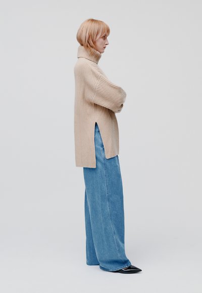 Beige geribbelde coltrui met zijsplitsen, gecombineerd met losse blauwe denim jeans en puntige zwarte schoenen, weergegeven vanuit de zijkant.