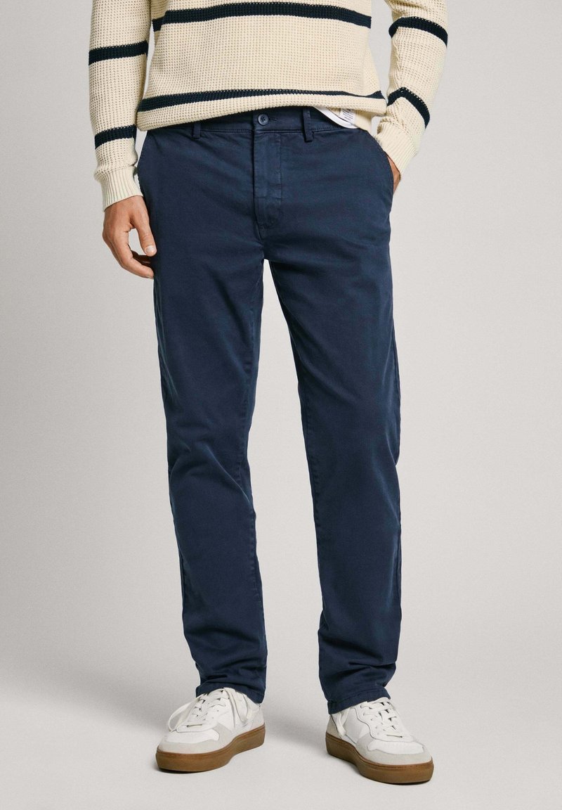 Homme portant un pantalon bleu marine, des baskets blanches et un pull crème avec des rayures horizontales noires, mains dans les poches, debout sur un fond uni.