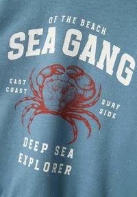 Maglietta in cotone blu con una grafica di un granchio rosso. Il testo recita "SEA GANG" e "DEEP SEA EXPLORER", con un aspetto vintage e una texture macchiata.