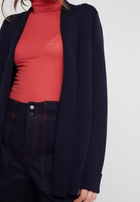 Cárdigan azul marino sobre un cuello de tortuga rojo acanalado, combinado con pantalones oscuros de alta cintura con costuras rojas y detalles de botones. Textura de tela suave.