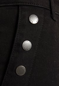 Tecido de denim preto com três botões prateados apresentando gravações da marca. A textura é suave com um leve brilho, realçando a costura.