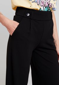 Černé kalhoty s hladkou texturou, s jedním knoflíkem na zapínání, bočními kapsami a jemným nařízením vpředu.