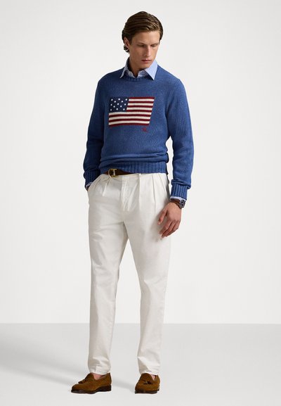 Blue knit sweater with a flag graphic, cream pants, and brown suede shoes. The sweater features ribbed cuffs and hem, worn over a collared shirt.