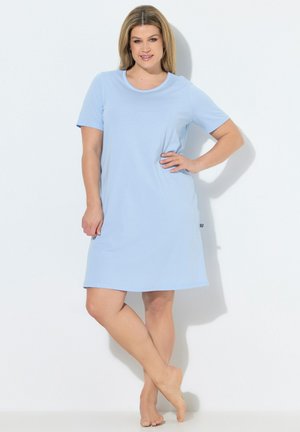 Kvinde, der står barfodet iført en lyseblå kortærmet kjole til knæet med den ene hånd på hoften mod en ensfarvet hvid baggrund.