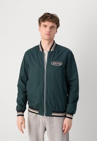Chaqueta bomber verde oscura con cuello y puños acanalados en navy y blanco a rayas, con cremallera frontal y parche con logo en el lado izquierdo del pecho.