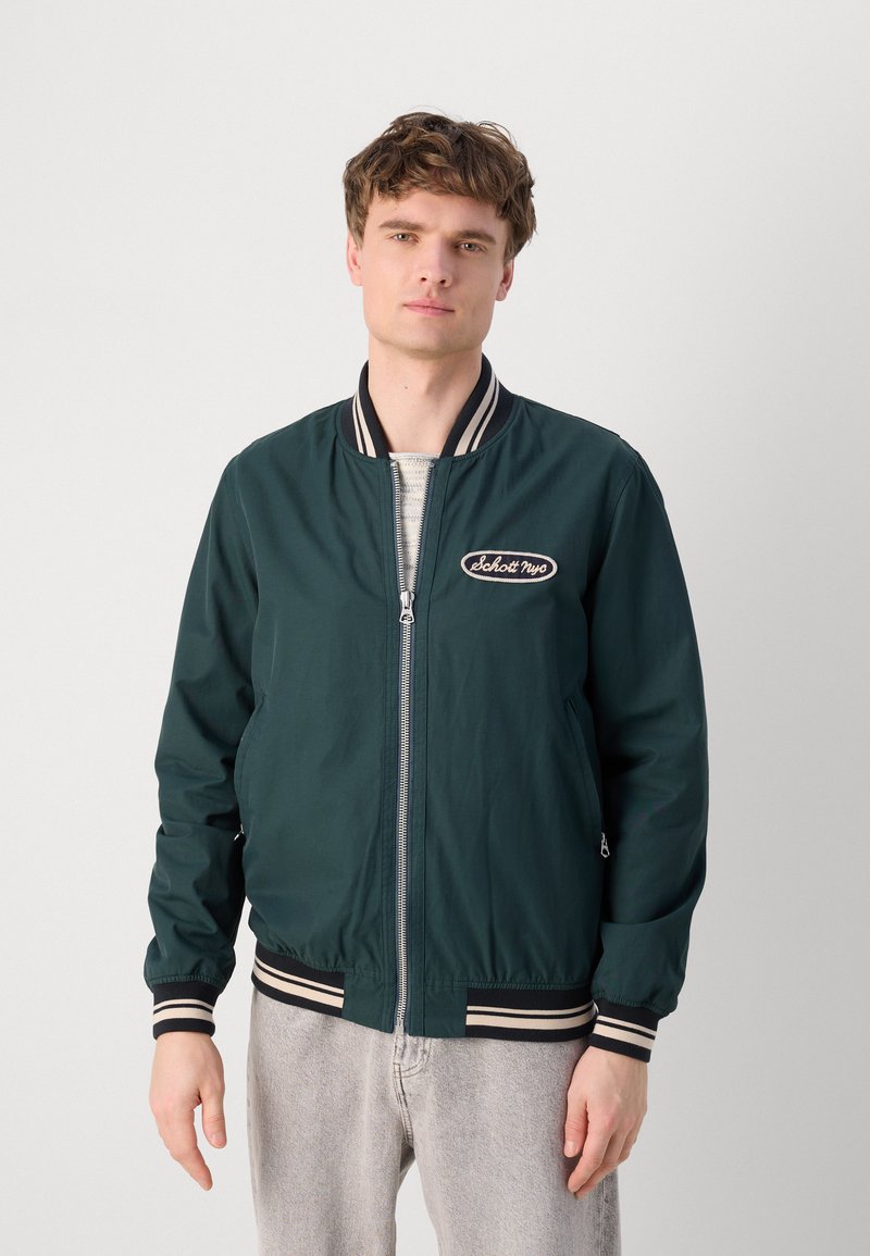 Veste bomber vert foncé avec col et poignets côtelés rayés bleu marine et blanc, dotée d'une fermeture éclair à l'avant et d'un patch logo sur la poitrine gauche.