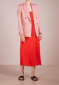 Blazer rose clair à double boutonnage avec boutons dorés, associé à une robe sans manches rouge vif et des sandales plates noires tissées. Tissu lisse.