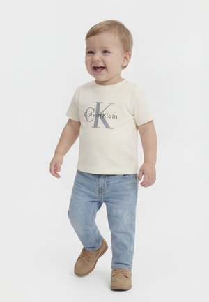 Småtroll med lysebrunt hår iført en cremefarvet Calvin Klein t-shirt, lyseblå jeans og tanfarvede sko, smilende og gående mod en hvid baggrund.