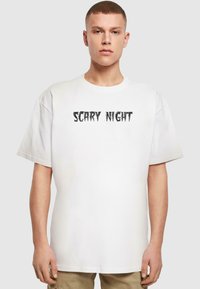 Hvid bomulds t-shirt med en iøjnefaldende sort teksttryk "SCARY NIGHT" i en takkete, stiliseret skrifttype. Løs, oversized pasform med korte ærmer.