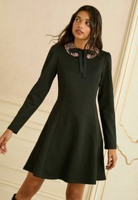 Čierne šaty s dlhým rukávom, s priliehavým živôtikom a rozšírenou sukňou. Majú golier s leopardím vzorom a viazanku na krku. Vyrobené z hladkej látky.