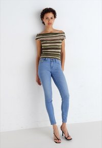 Camiseta de rayas verdes y beige, con diseño ajustado y hombros al descubierto, combinada con jeans skinny de un azul claro y tacones negros con tiras.