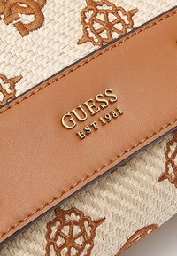 Bracelet en cuir fauve avec logo doré "GUESS EST 1981" sur tissu beige tissé décoré de motifs brodés en forme d'étoiles marron.