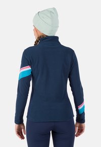 Námořnická fleecová mikina s vysokým límcem, s tyrkysovým a růžovým pruhem přes levou rukáv. Nosená s světle zelenou čepicí.