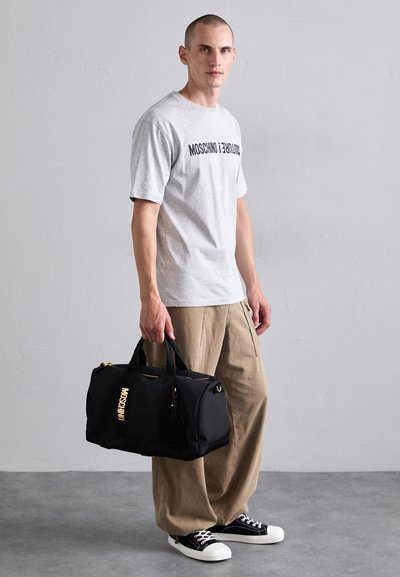 Gray cotton t-shirt with "MOSCHINO" print, khaki wide-leg pants, and black fabric duffle bag featuring gold accents and handles. Black sneakers.
