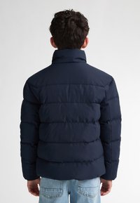 Giubbotto imbottito blu navy con colletto alto e sezioni trapuntate orizzontali, che offrono un aspetto testurizzato e isolato, abbinato a jeans chiari.