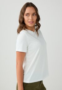 Vrouw met golvend bruin haar, gekleed in een effen witte korte mouwen t-shirt en olijfgroene broek, kijkt naar links tegen een effen achtergrond.