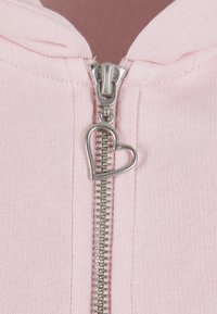 Lyserød lynlåsjakke med en sølvfarvet metallynlås, der har en hjerteformet lynlåstrækker. Stoffet har et blødt, tekstureret udseende.