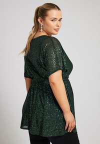 Tunic verde con paillettes, maniche corte, vita a cintura e vestibilità ampia. Superficie testurizzata con luccichio, abbinata a pantaloni neri.