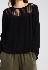 Haut noir à manches longues avec des détails en dentelle élaborée sur les épaules et l'ourlet. Fabriqué en tissu texturé, il présente une coupe décontractée et un col rond.