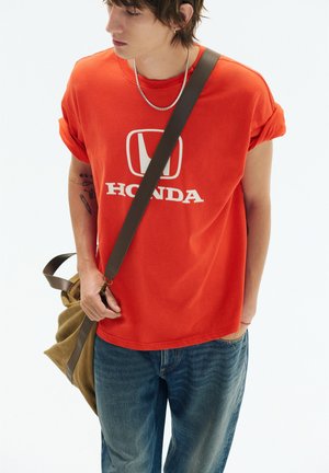 Giovane che indossa una maglietta arancione Honda e jeans, tiene una borsa verde con una tracolla marrone sulla spalla, guardando di lato.