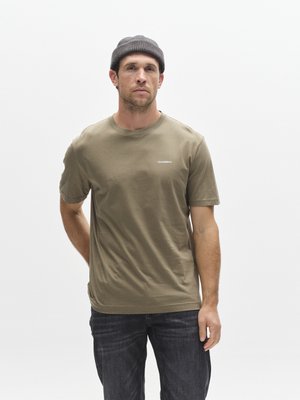 Bombažno majico maslinasto zelene barve s kratkimi rokavi in okroglim izrezom. Na prsih je majhna bela značka. Kombinirana s temnimi kavbojkami.