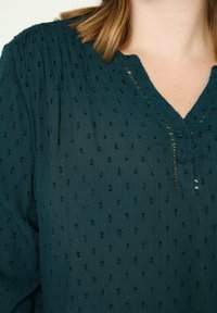 Blouse bleu sarcelle foncé en tissu texturé, avec un col en V, de petits boutons et un motif ponctué subtil sur toute la surface.