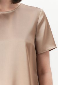 Close-up van een persoon in een gladde, beige korte mouwen shirt tegen een plain witte achtergrond.