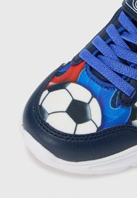 Laivastonsininen tennari, jossa on teksturoitu jalkapallokuvio punaisilla ja sinisillä yksityiskohdilla, valkoinen ulkopohja ja siniset nauhat.